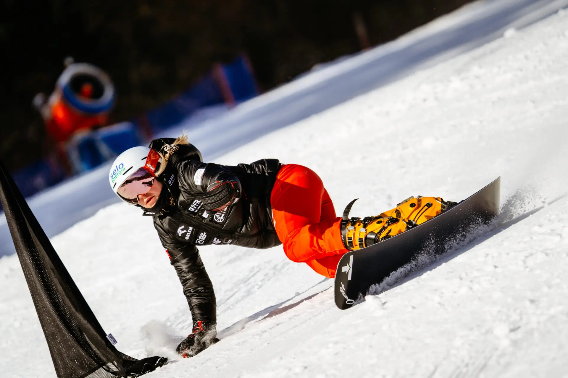 Polska snowboardzistka - Marysia Bukowska-Chyc - w czarnej kurtce i czerwonych spodniach pochyla się niemal równolegle do stoku, dotykając dłonią śniegu podczas szybkiego skrętu slalomowego.